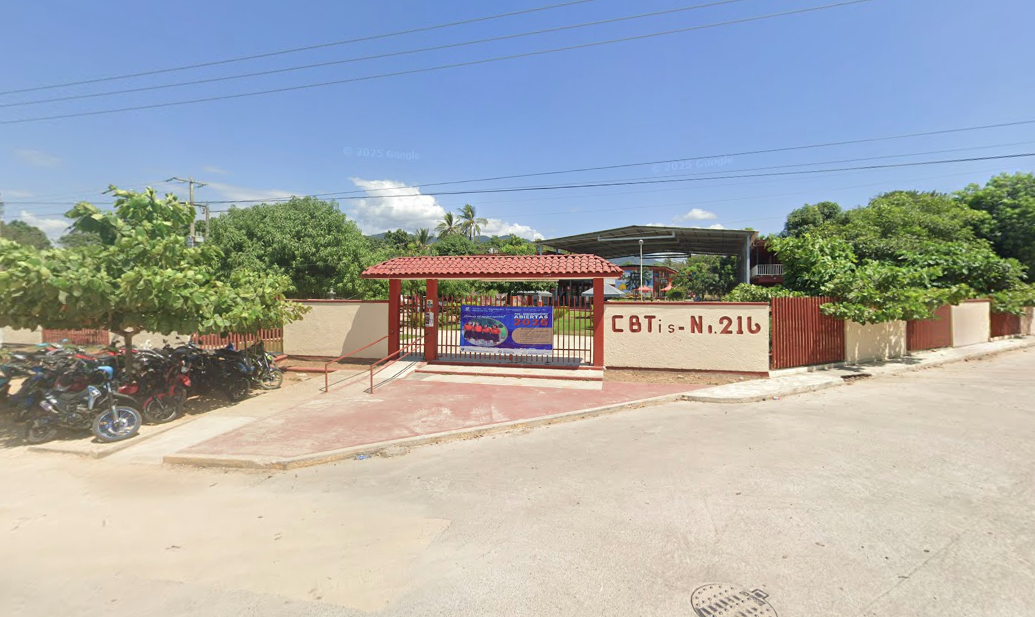 Fotografía en la que se muestra una pequeña explanada donde hay una rampa con un barandal que permite el acceso. Al terminar la explanada está la entrada al plantel, conformada por una reja techada de color rojo. A la izquierda y a la derecha de la rej<a hay unos muros de color amarillo y en el muro de la derecha está escrito en letras grandes rojas Cebetis número 216.